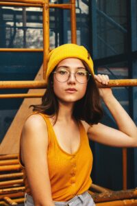 Stylish woman posing in mustard beanie and glasses against industrial backdrop.